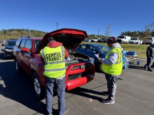 University of Georgia Campus Transit employees stand in front of a red Toyota SUV with its hood up while they refill wiper fluid and hold a checklist. They are in a parking lot on the UGA campus at the Operation Safe Drive event.